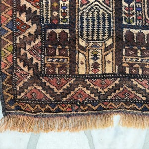May include: A woven rug featuring a dark brown, tan, and deep red geometric pattern with intricate architectural motifs. The border displays a repeating zigzag design, and the bottom edge is finished with a light tan fringe of knotted yarn.
