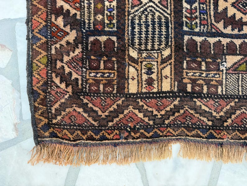 May include: A woven rug featuring a dark brown, tan, and deep red geometric pattern with intricate architectural motifs. The border displays a repeating zigzag design, and the bottom edge is finished with a light tan fringe of knotted yarn.
