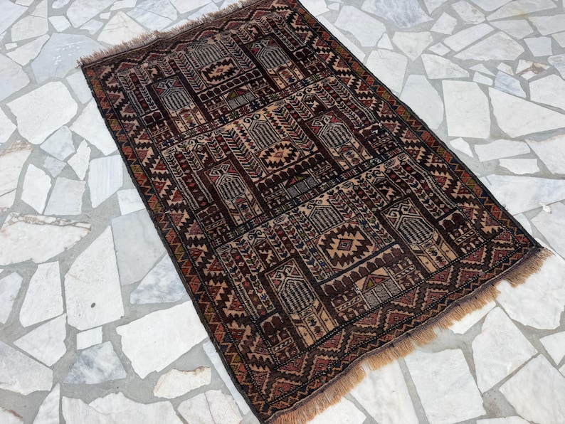 May include: A rectangular woven rug featuring a dark brown, black, and tan geometric pattern with repeating arch motifs and intricate borders. The rug has a short fringe on the top and bottom edges and a dense, symmetrical design throughout.