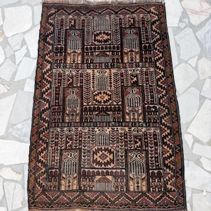 May include: A rectangular woven rug featuring a complex geometric pattern in shades of dark brown, tan, red, and blue. The design includes repeating architectural motifs, stylized floral elements, and a fringed edge on the top and bottom ends.