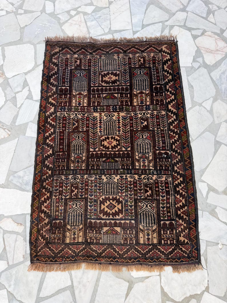 May include: A rectangular woven rug featuring a complex geometric pattern in shades of dark brown, tan, red, and blue. The design includes repeating architectural motifs, stylized floral elements, and a fringed edge on the top and bottom ends.