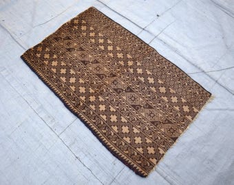 May include: A rectangular, brown rug with a detailed geometric pattern. The rug features a repeating design of diamonds, stars, and floral motifs in shades of brown and tan. The edges are finished with a dark brown border.