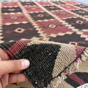 K&ouml;nnte beinhalten: Ein gewebter Teppich mit einem sich wiederholenden geometrischen Rautenmuster in den Farben Dunkelbraun, ged&auml;mpftes Rot und Creme. Der Teppich hat einen dicken, strukturierten Flor mit sichtbarer Faserstruktur und Fransen am umgeklappten Eck.
