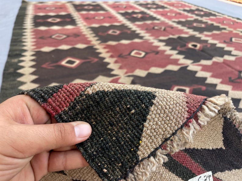 K&ouml;nnte beinhalten: Ein gewebter Teppich mit einem sich wiederholenden geometrischen Rautenmuster in den Farben Dunkelbraun, ged&auml;mpftes Rot und Creme. Der Teppich hat einen dicken, strukturierten Flor mit sichtbarer Faserstruktur und Fransen am umgeklappten Eck.