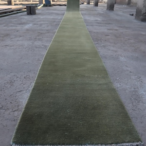 May include: A long, olive green runner rug with a textured surface. The rug is rectangular and has a fringe on one end. The rug is displayed outdoors on a concrete surface.