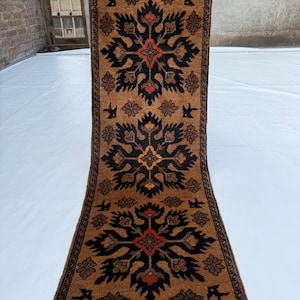 Puede incluir: Una alfombra pasillera rectangular con fondo marrón y tres motivos florales negros y naranjas repetidos. La alfombra tiene un borde marrón oscuro con pequeños patrones geométricos y flecos grises en ambos extremos, de textura anudada a mano.