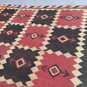 K&ouml;nnte beinhalten: Ein gewebter Teppich mit einem sich wiederholenden geometrischen Muster aus ineinandergreifenden Rauten in den Farben Dunkelbraun, ged&auml;mpftes Rot und Creme. Das Design enth&auml;lt kleine rautenf&ouml;rmige Motive mit pfeilartigen Akzenten.