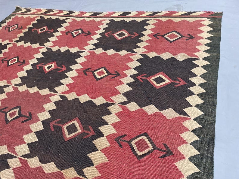 K&ouml;nnte beinhalten: Ein gewebter Teppich mit einem sich wiederholenden geometrischen Muster aus ineinandergreifenden Rauten in den Farben Dunkelbraun, ged&auml;mpftes Rot und Creme. Das Design enth&auml;lt kleine rautenf&ouml;rmige Motive mit pfeilartigen Akzenten.
