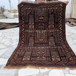 May include: A rectangular woven rug featuring a dark brown and tan geometric pattern with multiple arched mihrab designs. The rug has a dense, intricate border with repeating triangular motifs and a short, light brown fringe along the bottom edge.