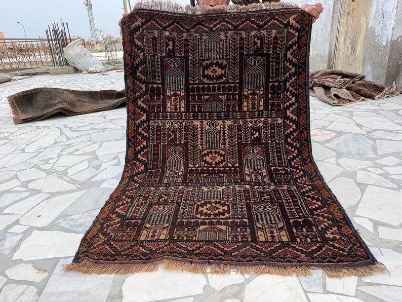 May include: A rectangular woven rug featuring a dark brown and tan geometric pattern with multiple arched mihrab designs. The rug has a dense, intricate border with repeating triangular motifs and a short, light brown fringe along the bottom edge.