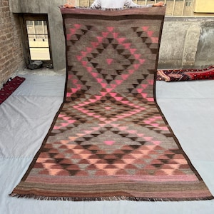 Op de afbeelding: Een rechthoekig tapijt met een geometrisch patroon in bruin-, roze- en grijstinten. Het tapijt heeft een centraal ruitontwerp met herhaalde driehoekvormen. De randen zijn afgezet met donkerder bruin en hebben een gefranjerde afwerking.