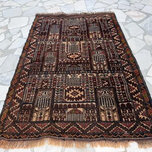 May include: A rectangular woven rug featuring a dark brown, tan, and deep red geometric pattern. The design includes repeating architectural motifs, stylized floral elements, and a central diamond shape, finished with short fringe on both ends.