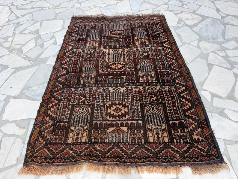 May include: A rectangular woven rug featuring a dark brown, tan, and deep red geometric pattern. The design includes repeating architectural motifs, stylized floral elements, and a central diamond shape, finished with short fringe on both ends.