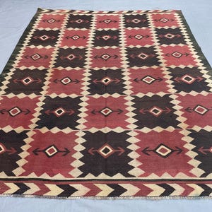 K&ouml;nnte beinhalten: Ein rechteckiger Teppich mit einem sich wiederholenden Gittermuster aus dunkelbraunen und tiefroten Quadraten. Jedes Quadrat enth&auml;lt eine zentrale Rautenform mit pfeilartigen Motiven in Wei&szlig;, Schwarz und Rot. Die Bord&uuml;re zeigt ein Zickzackmuster.