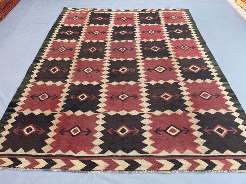 K&ouml;nnte beinhalten: Ein rechteckiger Teppich mit einem sich wiederholenden Gittermuster aus dunkelbraunen und tiefroten Quadraten. Jedes Quadrat enth&auml;lt eine zentrale Rautenform mit pfeilartigen Motiven in Wei&szlig;, Schwarz und Rot. Die Bord&uuml;re zeigt ein Zickzackmuster.