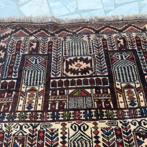May include: A rectangular woven rug featuring a complex geometric pattern in shades of dark brown, cream, red, blue, and green. The design includes repeating architectural motifs, stylized floral elements, and a fringed edge along the top border.