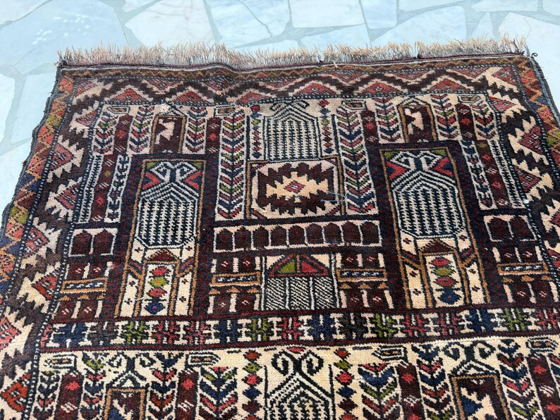 May include: A rectangular woven rug featuring a complex geometric pattern in shades of dark brown, cream, red, blue, and green. The design includes repeating architectural motifs, stylized floral elements, and a fringed edge along the top border.