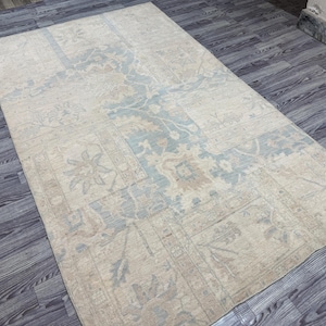 Könnte beinhalten: Ein rechteckiger, beigefarbener Teppich mit einem verwitterten, Vintage-Aussehen. Der Teppich hat ein zentrales Design in Blau- und Beigetönen mit floralen und geometrischen Mustern. Der Teppich liegt auf einem dunklen Holzboden.