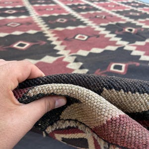 K&ouml;nnte beinhalten: Ein gewebter Teppich mit einem sich wiederholenden geometrischen Rautenmuster in den Farben Dunkelbraun, ged&auml;mpftes Rot und Creme. Das Textil weist einen dicken, strukturierten Flor mit sichtbaren verflochtenen Fasern auf.