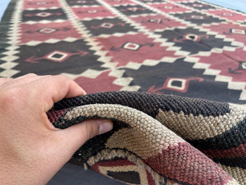 K&ouml;nnte beinhalten: Ein gewebter Teppich mit einem sich wiederholenden geometrischen Rautenmuster in den Farben Dunkelbraun, ged&auml;mpftes Rot und Creme. Das Textil weist einen dicken, strukturierten Flor mit sichtbaren verflochtenen Fasern auf.