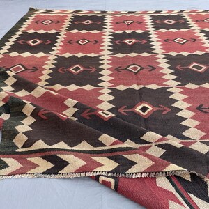 K&ouml;nnte beinhalten: Ein flach gewebter Teppich mit einem sich wiederholenden geometrischen Muster in Dunkelbraun, ged&auml;mpftem Rot und Creme. Das Design besteht aus abwechselnden Quadraten mit Rautenmotiven und pfeilartigen Akzenten, abgeschlossen durch eine Zickzack-Borte.