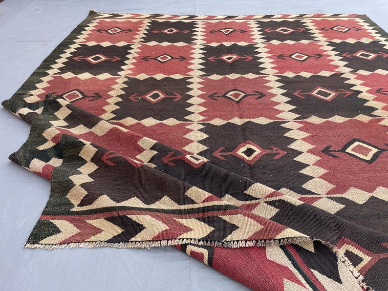 K&ouml;nnte beinhalten: Ein flach gewebter Teppich mit einem sich wiederholenden geometrischen Muster in Dunkelbraun, ged&auml;mpftem Rot und Creme. Das Design besteht aus abwechselnden Quadraten mit Rautenmotiven und pfeilartigen Akzenten, abgeschlossen durch eine Zickzack-Borte.