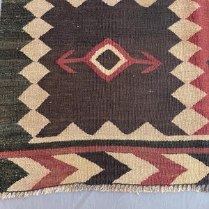 K&ouml;nnte beinhalten: Ein gewebter Teppich mit geometrischem Muster, einem dunkelbraunen Mittelfeld, cremefarbenen Zickzack-Bord&uuml;ren und roten Akzenten. Das Design umfasst Rautenformen und Pfeilmotive, abgeschlossen mit einer kurzen, hellen Franse an der Unterkante.