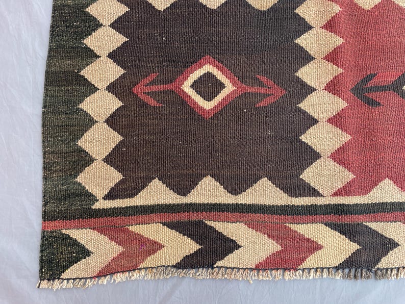 K&ouml;nnte beinhalten: Ein gewebter Teppich mit geometrischem Muster, einem dunkelbraunen Mittelfeld, cremefarbenen Zickzack-Bord&uuml;ren und roten Akzenten. Das Design umfasst Rautenformen und Pfeilmotive, abgeschlossen mit einer kurzen, hellen Franse an der Unterkante.