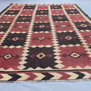 K&ouml;nnte beinhalten: Ein rechteckiger Teppich mit einem sich wiederholenden geometrischen Muster aus dunkelbraunen und tiefroten Quadraten, die durch gezackte cremefarbene R&auml;nder getrennt sind. Jedes Quadrat enth&auml;lt ein zentrales Rautenmotiv mit pfeilartigen Akzenten.