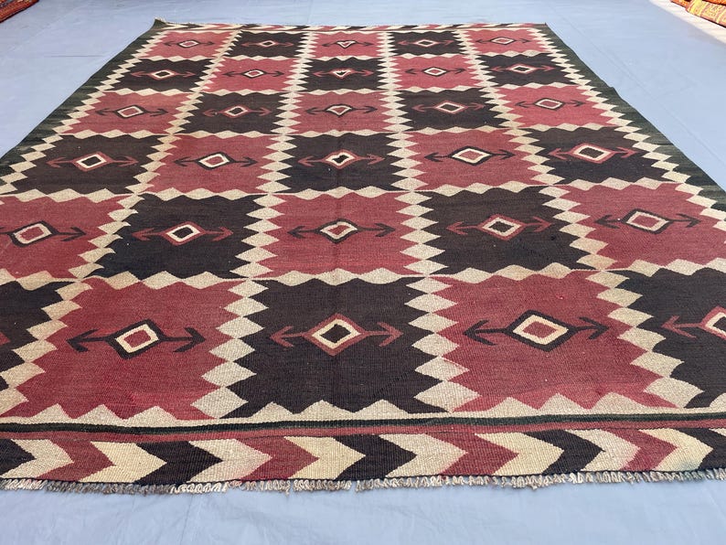 K&ouml;nnte beinhalten: Ein rechteckiger Teppich mit einem sich wiederholenden geometrischen Muster aus dunkelbraunen und tiefroten Quadraten, die durch gezackte cremefarbene R&auml;nder getrennt sind. Jedes Quadrat enth&auml;lt ein zentrales Rautenmotiv mit pfeilartigen Akzenten.