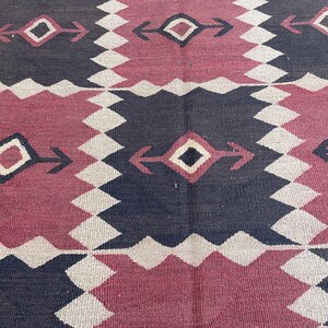 K&ouml;nnte beinhalten: Ein gewebter Teppich mit einem sich wiederholenden geometrischen Muster aus abwechselnden dunkelanthrazitfarbenen und tiefroten Quadraten. Jedes Quadrat enth&auml;lt eine zentrale Rautenform mit pfeilartigen Erweiterungen, umrandet von einem hellen Zickzackmuster.