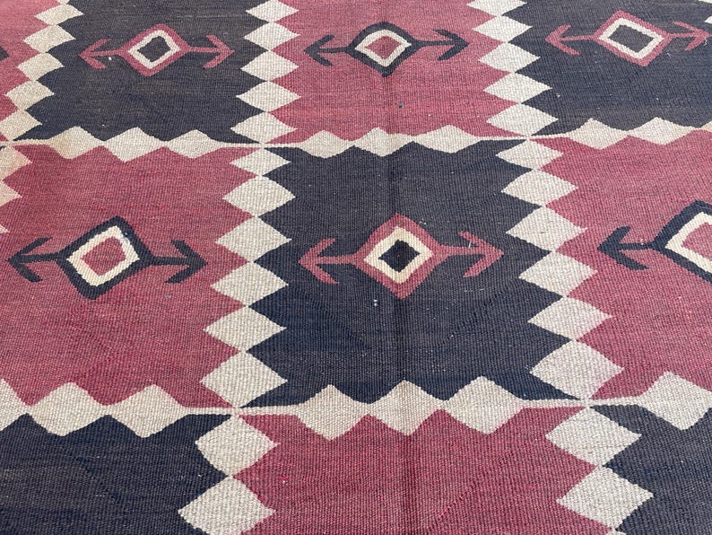 K&ouml;nnte beinhalten: Ein gewebter Teppich mit einem sich wiederholenden geometrischen Muster aus abwechselnden dunkelanthrazitfarbenen und tiefroten Quadraten. Jedes Quadrat enth&auml;lt eine zentrale Rautenform mit pfeilartigen Erweiterungen, umrandet von einem hellen Zickzackmuster.