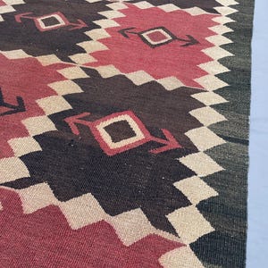 K&ouml;nnte beinhalten: Ein gewebter Teppich mit einem sich wiederholenden geometrischen Muster aus gro&szlig;en Rauten in tiefem Rot und dunklem Anthrazitgrau. Die Formen sind mit einem cremefarbenen Zickzack-Rand versehen und enthalten ein kleines Quadrat mit Pfeilakzenten.