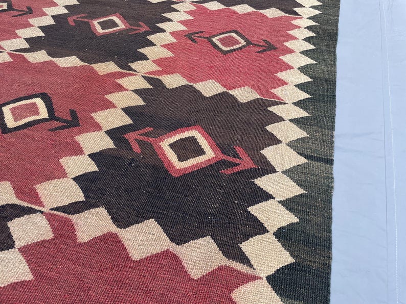 K&ouml;nnte beinhalten: Ein gewebter Teppich mit einem sich wiederholenden geometrischen Muster aus gro&szlig;en Rauten in tiefem Rot und dunklem Anthrazitgrau. Die Formen sind mit einem cremefarbenen Zickzack-Rand versehen und enthalten ein kleines Quadrat mit Pfeilakzenten.