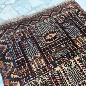 May include: A rectangular woven rug featuring a complex geometric pattern in shades of dark brown, cream, red, blue, and green. The design includes repeating architectural motifs and intricate borders, finished with a short fringe along the edge.