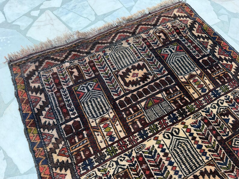 May include: A rectangular woven rug featuring a complex geometric pattern in shades of dark brown, cream, red, blue, and green. The design includes repeating architectural motifs and intricate borders, finished with a short fringe along the edge.
