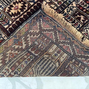 May include: A hand-knotted rug featuring a complex geometric pattern in shades of deep red, navy blue, cream, and brown. The rug is folded over to reveal the underside, showcasing a dense weave and a light tan fringe along the edge of the fabric.