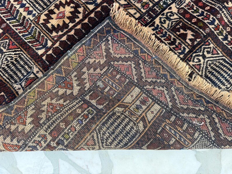 May include: A hand-knotted rug featuring a complex geometric pattern in shades of deep red, navy blue, cream, and brown. The rug is folded over to reveal the underside, showcasing a dense weave and a light tan fringe along the edge of the fabric.