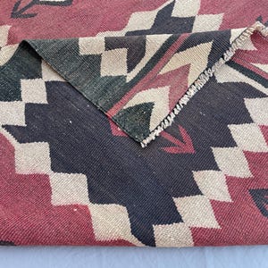 K&ouml;nnte beinhalten: Ein gewebter Teppich mit einem geometrischen Muster aus sich wiederholenden Rauten und Zickzacklinien in Dunkelrot, Anthrazit und Creme. Der Stoff weist eine sichtbare Flachgewebestruktur auf, wobei eine umgeschlagene Ecke die R&uuml;ckseite zeigt.