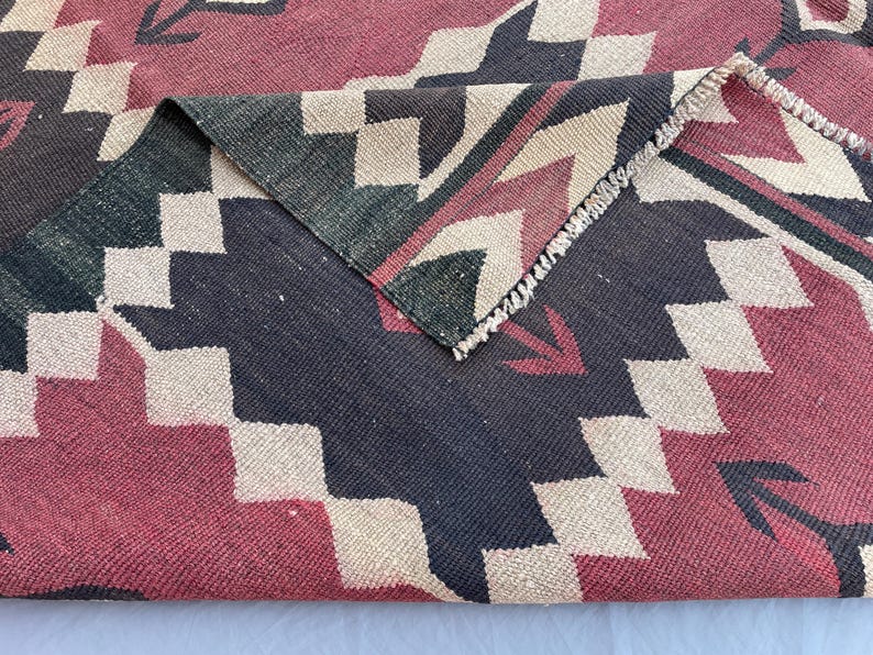 K&ouml;nnte beinhalten: Ein gewebter Teppich mit einem geometrischen Muster aus sich wiederholenden Rauten und Zickzacklinien in Dunkelrot, Anthrazit und Creme. Der Stoff weist eine sichtbare Flachgewebestruktur auf, wobei eine umgeschlagene Ecke die R&uuml;ckseite zeigt.