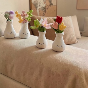 Può includere: Quattro vasi di peluche bianchi con facce sorridenti, ognuno con una diversa composizione di fiori di stoffa colorati. I fiori includono fiori rosa, gialli, verdi e rossi, creando un'esposizione allegra e decorativa.