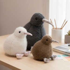 Puede incluir: Tres peluches de kiwi en blanco, gris y marrón, cada uno con un pico largo y patas pequeñas. Los pájaros están sobre una superficie de madera clara. Un pequeño cactus, lápices y un cuaderno están en el fondo.