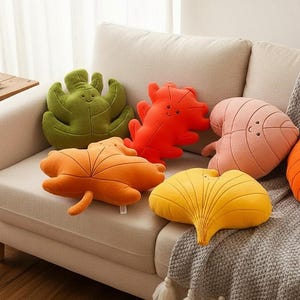 May include: A selection of leaf-shaped cushions in shades of green, orange, yellow, and pink, placed on a beige sofa. The cushions feature embroidered details and are designed to resemble different types of leaves.
