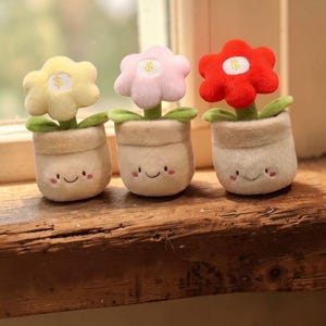 Könnte beinhalten: Drei Plüschblumen-Spielzeuge in Töpfen. Jede Blume hat eine andere Farbe: gelb, rosa und rot. Die Töpfe sind beige mit lächelnden Gesichtern und grünen Blättern. Die Blumen haben ein Dollarzeichen in der Mitte.
