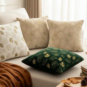 Può includere: Tre cuscini quadrati decorativi con un motivo a foglie ripetuto. Un cuscino è bianco con foglie dorate, uno è beige con foglie dorate e uno è verde scuro con foglie dorate. I cuscini sono disposti su un divano di colore chiaro.