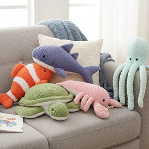 Pode incluir: Uma coleção de brinquedos de pelúcia de criaturas marinhas, incluindo um peixe-palhaço, um tubarão, uma tartaruga, uma lagosta e um polvo, estão dispostos em um sofá cinza. Os brinquedos são de várias cores, como laranja, azul, verde, rosa e azul claro. Um livro infantil está aberto.