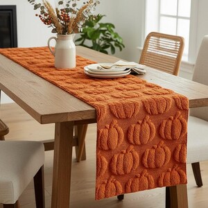 May include: A rectangular orange table runner featuring a repeating raised, textured pumpkin pattern across its entire surface. The fabric has a soft, plush appearance with a consistent burnt orange colour, draped over a wooden dining table.