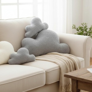May include: A cream-colored sofa with several cloud-shaped pillows in white and gray. A beige throw blanket is draped over the sofa. A wooden coffee table holds a mug and a book.