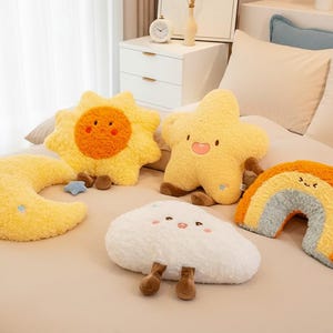 May include: A set of plush cushions shaped like a sun, star, moon, cloud, and rainbow. The sun and star are yellow, the moon is yellow, the cloud is white, and the rainbow is orange, yellow, and blue. Each cushion has a smiling face.