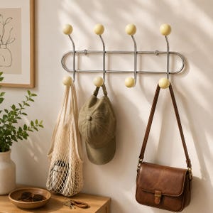 Peut inclure: Un porte-manteau mural composé d'une structure en métal argenté avec huit crochets, chacun surmonté d'une boule jaune pâle lisse. Un sac fourre-tout en filet beige, une casquette vert olive et un sac à bandoulière en cuir marron y sont suspendus.
